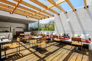 An outdoor dining area with wooden tables and chairs, a kitchen in the background, and a view of greenery. - Wonderful apt w/ balcony @Parque Mexico | ChiCo (Ciudad de México)