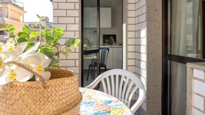 A balcony with a wicker basket of white orchids, a white plastic chair, and a glimpse of a kitchen area through a glass door. - Sardinia RE - Flavia s Flat - Free Parking (Alghero)