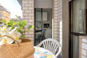 Un balcon orné d’un panier en osier rempli d’orchidées blanches, d’une chaise en plastique blanche, et d’où l’on aperçoit une partie de la cuisine à travers une porte vitrée.