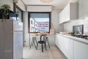A modern kitchen with a dining table, chairs, a refrigerator, a stove, and a window with a view of buildings. - Sardinia RE - Flavia s Flat - Free Parking (Alghero)