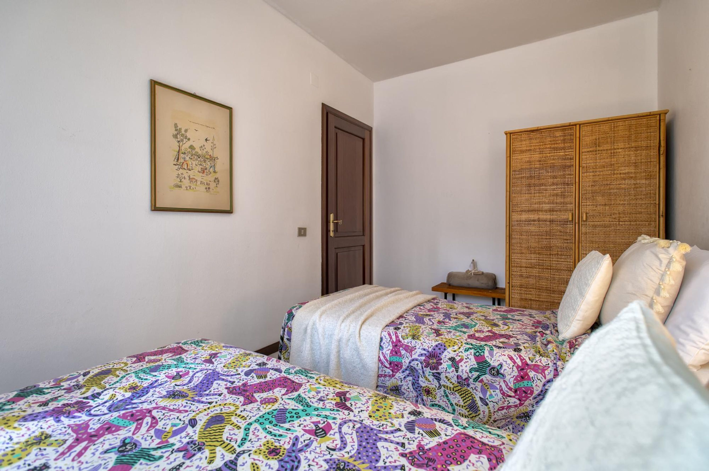 A bedroom with two beds, a wooden wardrobe, a framed picture on the wall, and a door.