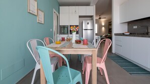 Une cuisine moderne avec des meubles blancs, un réfrigérateur en acier inoxydable et un coin repas comprenant une table en bois et des chaises colorées.