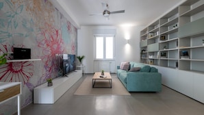 Un salon moderne avec un canapé de couleur turquoise, un téléviseur à écran plat, un ventilateur de plafond et une bibliothèque.