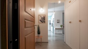 Un couloir avec une porte en bois, une petite table sur laquelle est posée une plante en pot, et une vue sur un coin cuisine.