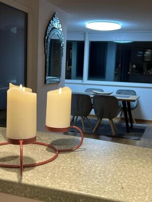 Een eetgedeelte met een tafel en stoelen, twee brandende kaarsen op een metalen standaard, en een spiegel aan de muur.