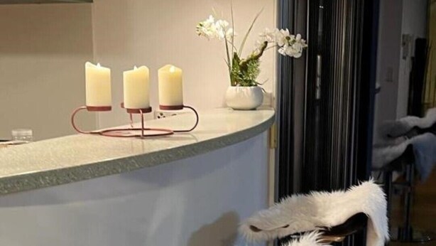 Eine Bar mit einem weißen, flauschigen Gegenstand auf einem Hocker, drei beleuchtete Kerzen auf einem Sockel und einer Vase mit weißen Blumen.