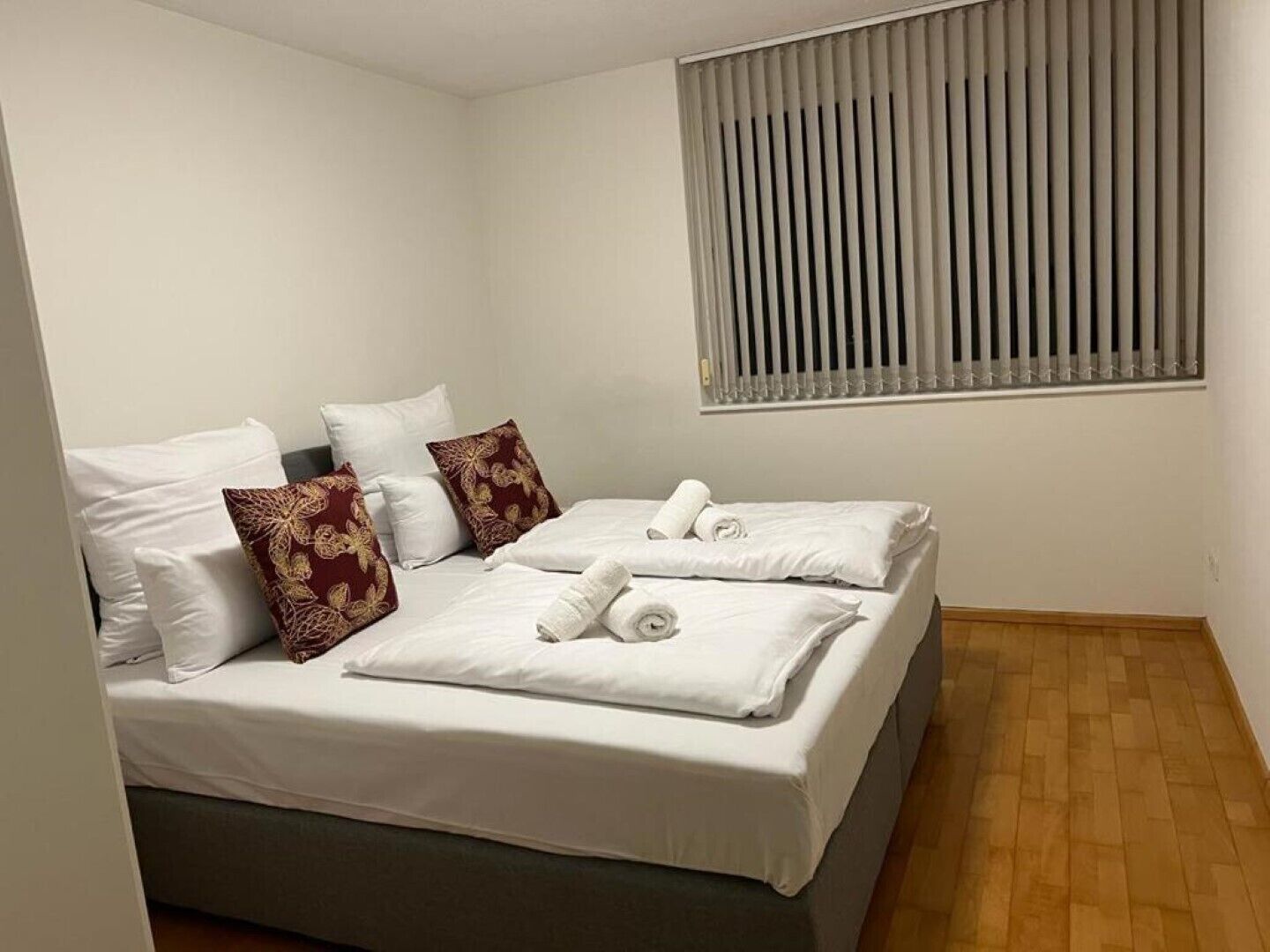 Un lit double bien fait, avec du linge de lit blanc et deux coussins décoratifs, une fenêtre avec des stores verticaux et un plancher en bois.