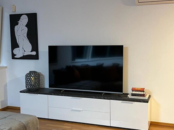 Een woonkamer met een grote flatscreen televisie op een witte kast, een zwarte vaas en een ingelijste foto aan de muur.