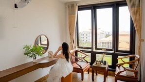 A room with a large window, a wooden chair, a small table, a potted plant, and a wall-mounted fan. - SeaColor Beachstay Danang Hotel Haviland (Da Nang)