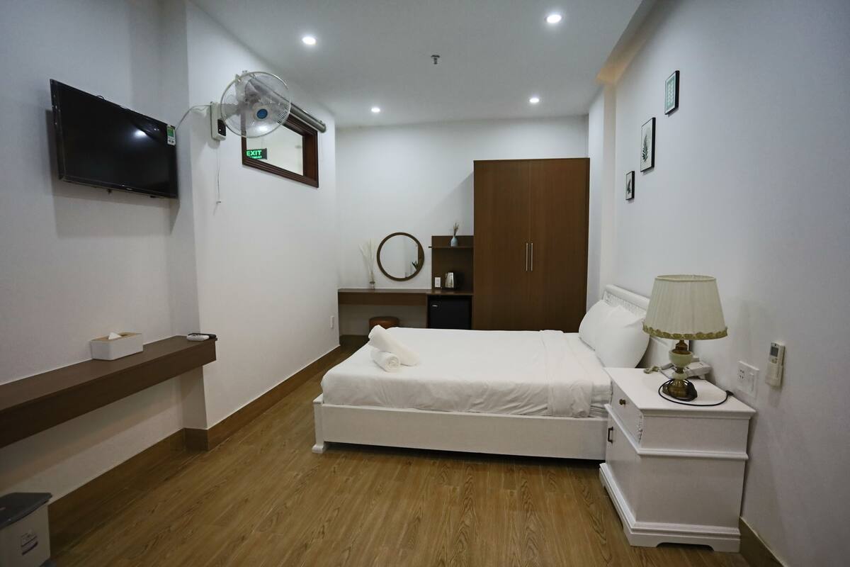 A hotel room with a bed, bedside table, lamp, TV, and a desk with a chair.