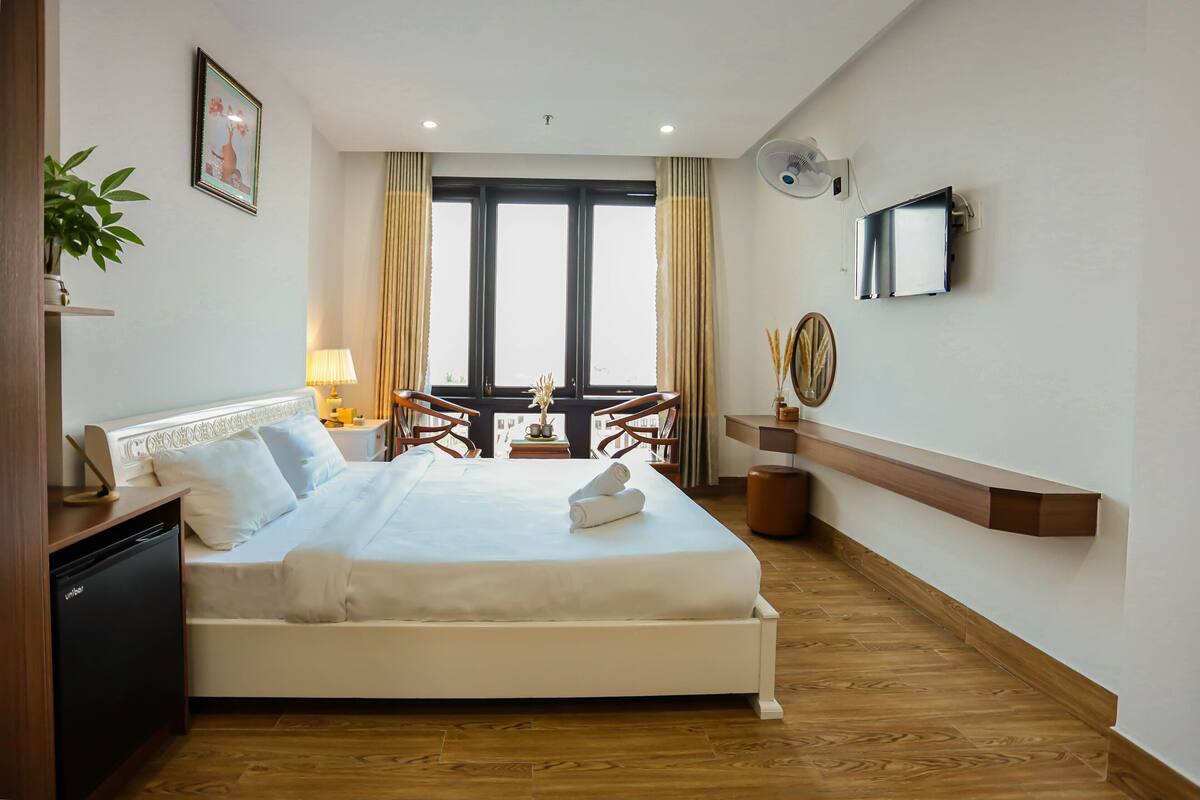 A hotel room with a bed, bedside table, lamp, plant, and a view of the outdoors through large windows.