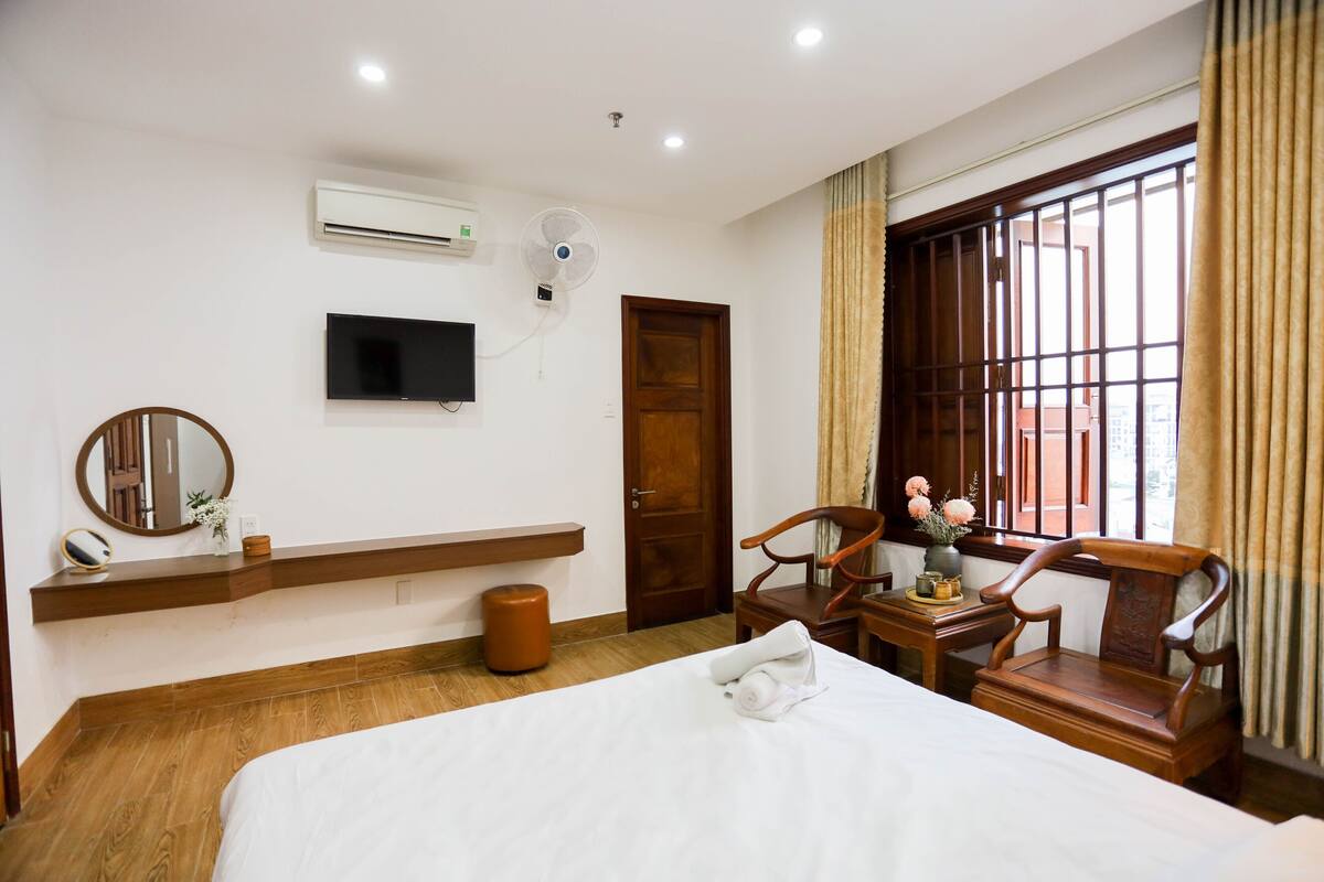 A hotel room with a bed, a TV, an air conditioner, a wooden chair, a small table, and a window with curtains.