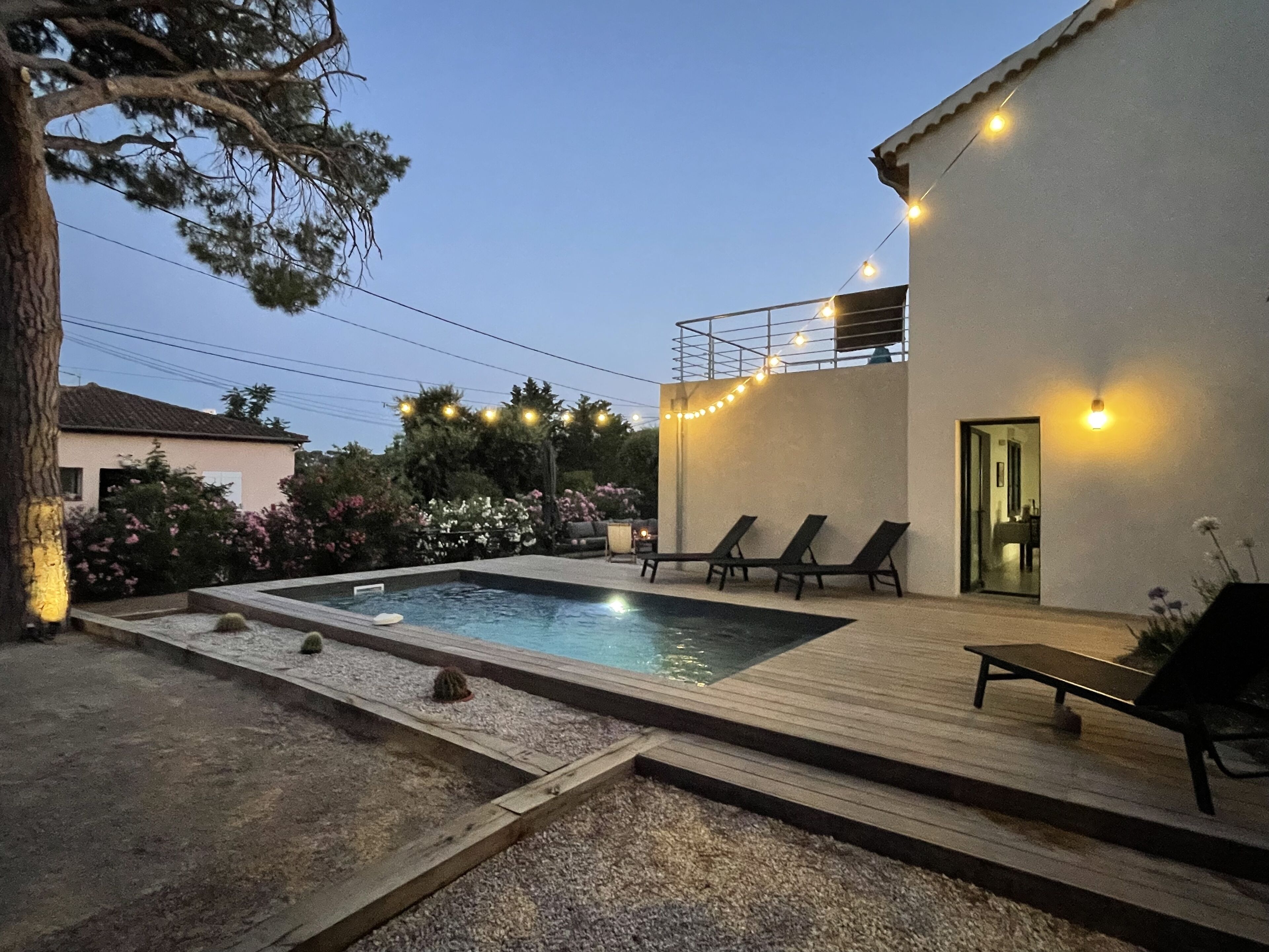 A swimming pool with lounge chairs and string lights, surrounded by a garden and a building.