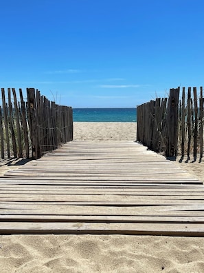 Ein hölzerner Steg führt zu einem Sandstrand unter blauem Himmel.