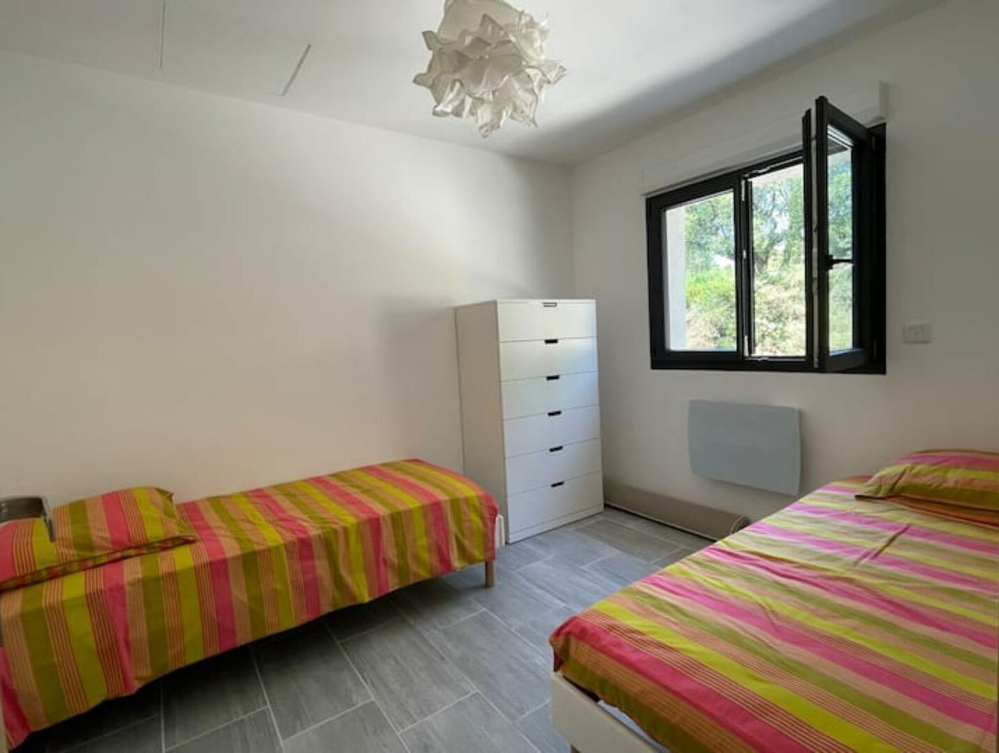 Un dormitorio con dos camas, un armario blanco, una ventana con vistas a zonas verdes y una lámpara en el techo.