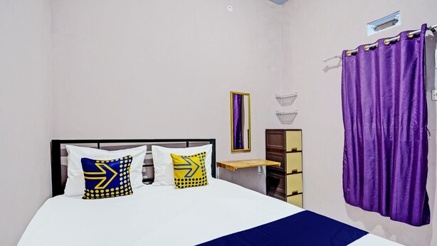A neatly made bed with a blue and yellow bedspread, two pillows with geometric patterns, a purple curtain, and a small shelf with a mirror and decorative items. - Hotel O Sobat Ijen HomestayNearSungai Prengsengan (Rogojampi)