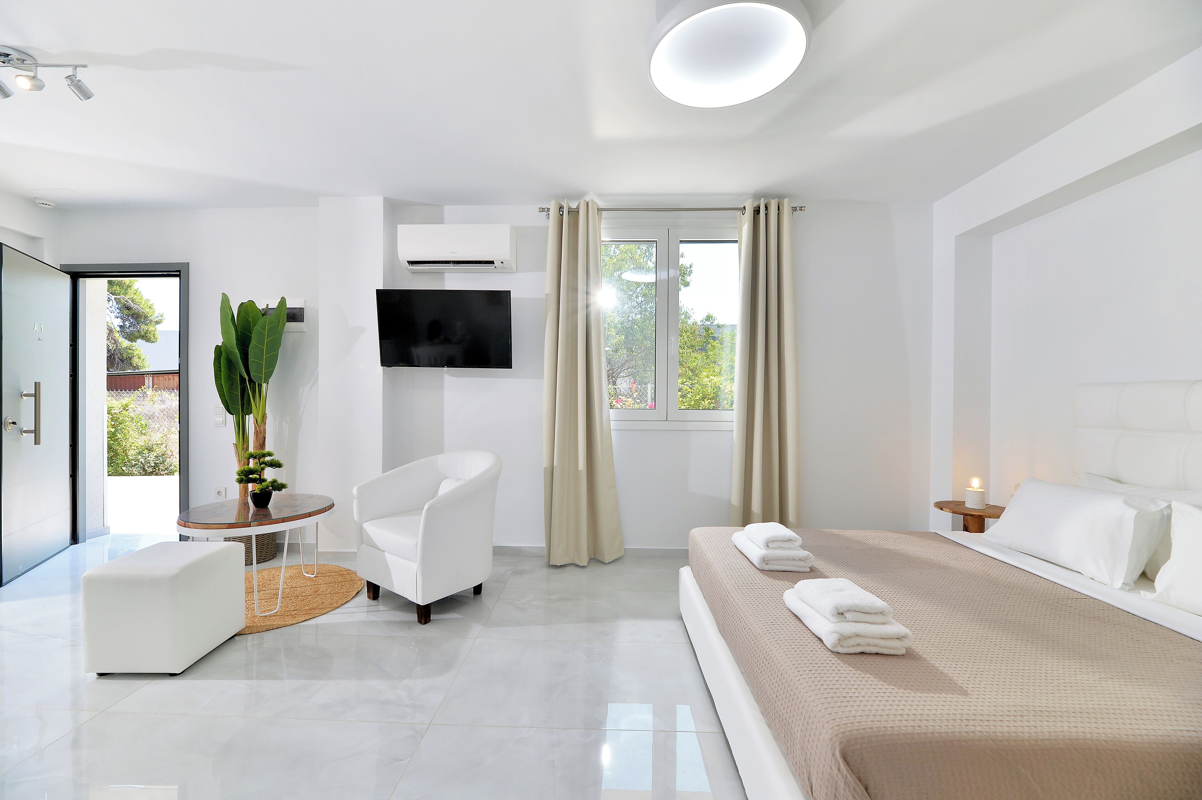 A modern hotel room with a large bed, a white armchair, a small round table, and a TV mounted on the wall.