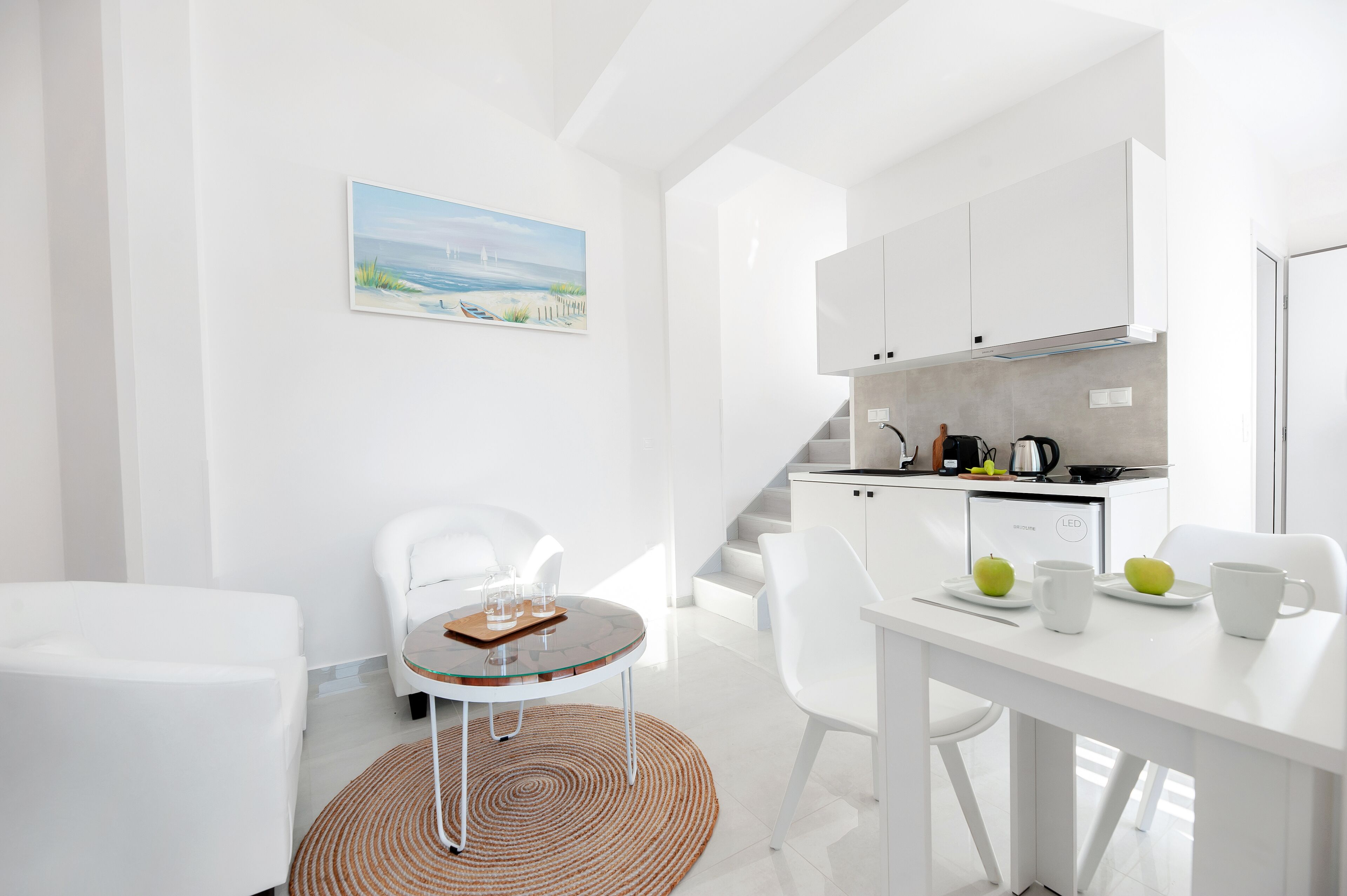 Una habitación moderna con decoración blanca, que incluye una mesa de comedor, sillas, una cocina y una escalera.