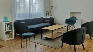 Un salon moderne avec un canapé, une table basse et deux fauteuils. On y trouve une bibliothèque et une fenêtre avec des rideaux.