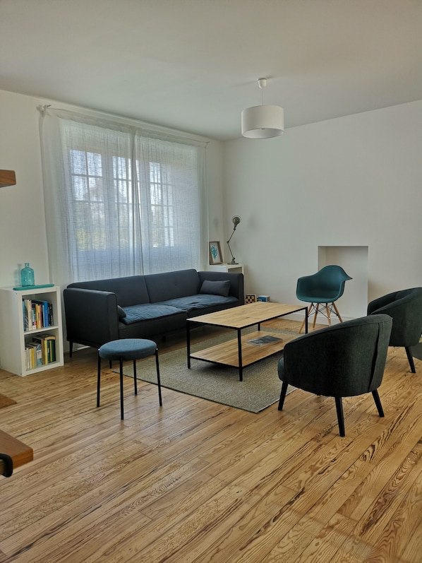 A modern living room with a sofa, a coffee table, and two chairs. There is a bookshelf and a window with curtains.