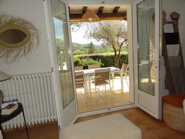 Une porte-fenêtre coulissante donne sur un patio extérieur aménagé avec une table et des chaises, un panier en osier et un radiateur à l’intérieur.