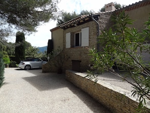 Un bâtiment en pierre avec une allée gravillonnée, une voiture garée et de la verdure tout autour.
