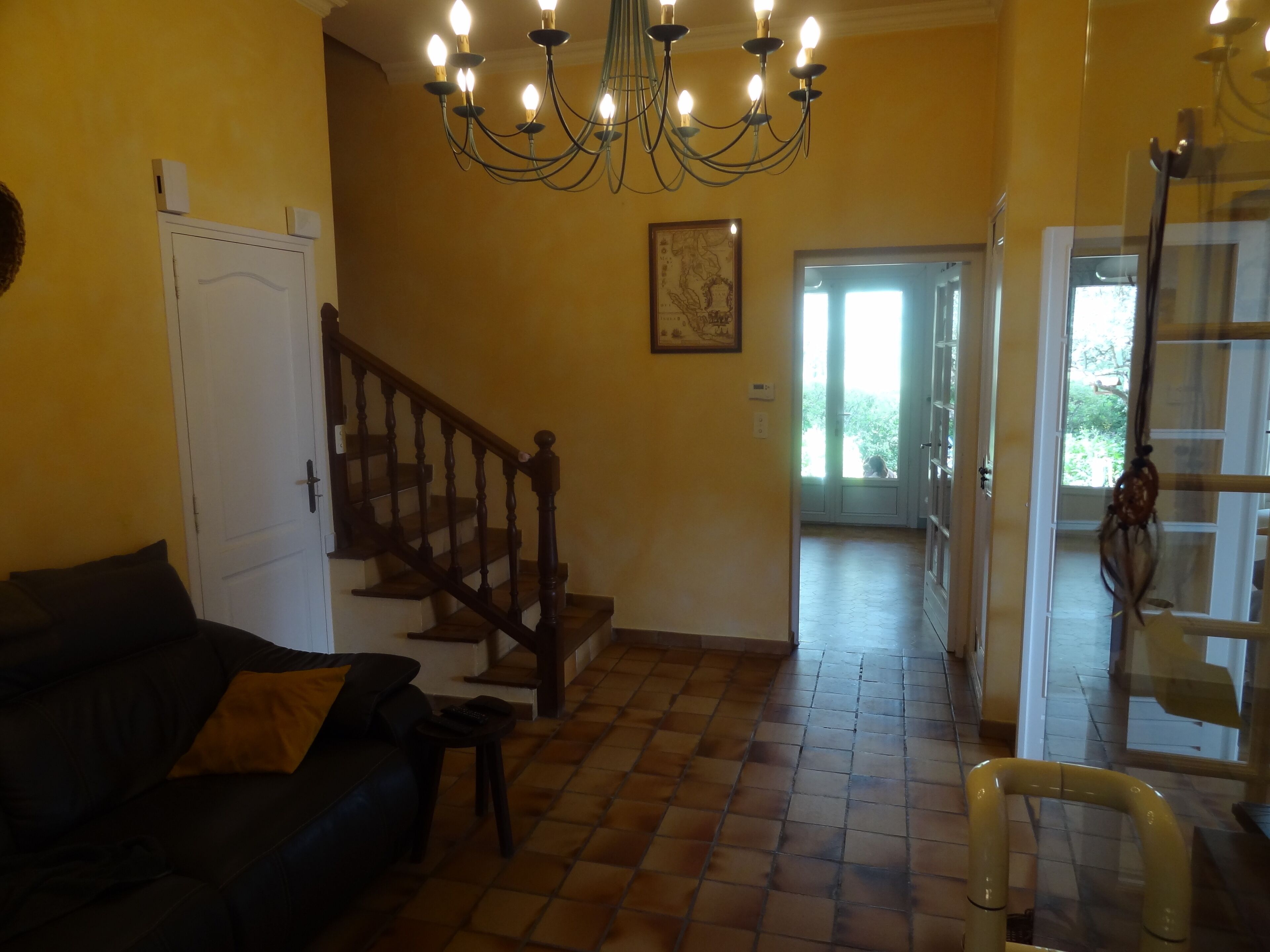 Una sala de estar con un sofá de color oscuro, una mesita, una araña, una escalera y una puerta que da a un área exterior luminosa.