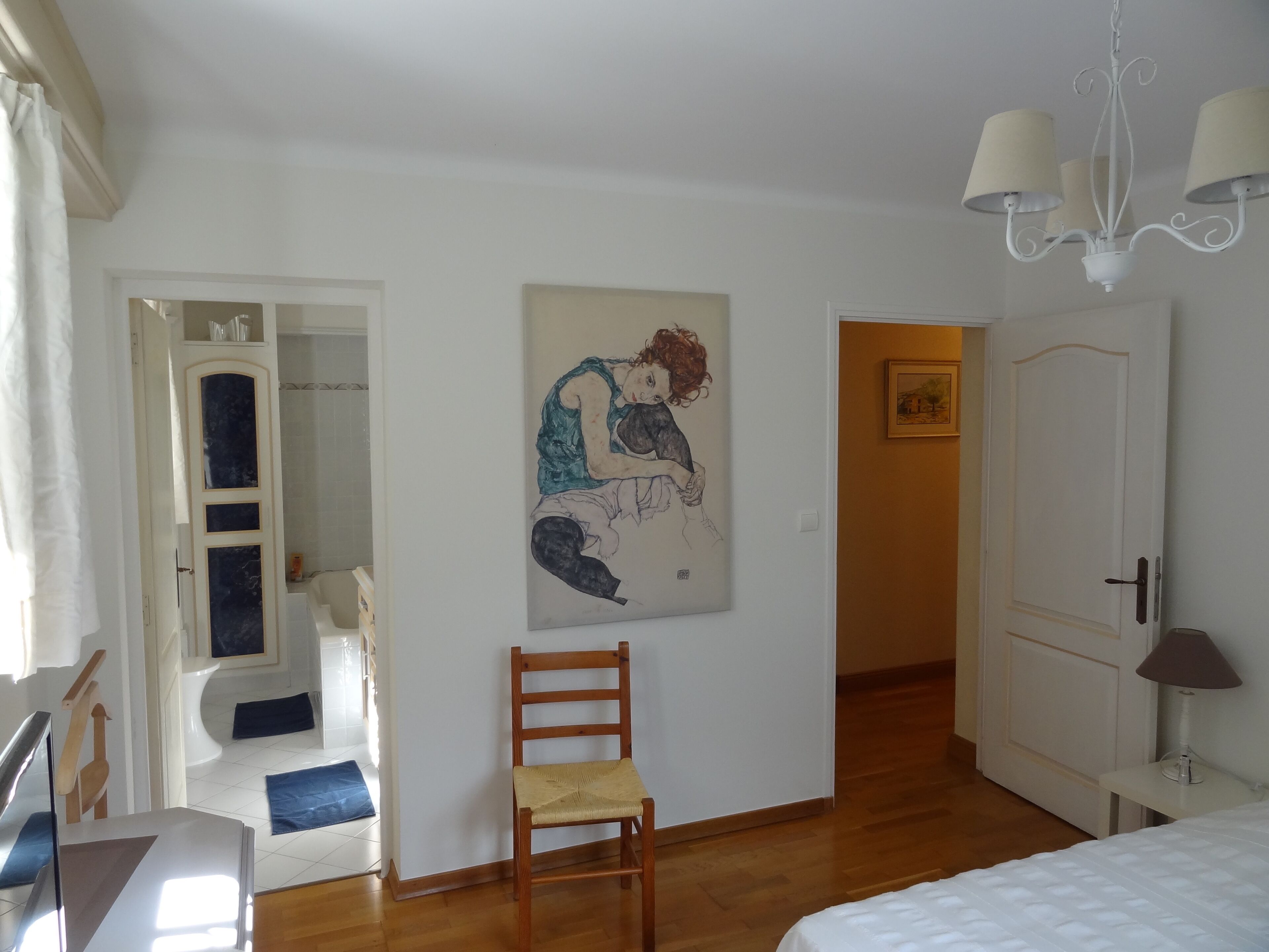 Un dormitorio con una cama, una silla de madera, un cuadro en la pared, una puerta y un pasillo.