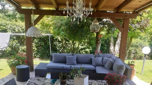 An outdoor patio with a wooden pergola, a round table with a floral tablecloth, and a grey sofa. - Gorgeous shabby rustic on Tuscan hills, pool on request (Capannori)