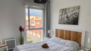 A bedroom with a large bed, a wooden headboard, a nightstand, a lamp, a window with a view of buildings, and a painting on the wall. - Air-conditioned T3 apartment for 6 with terrace and parking in Port Leucate (Port Leucate)