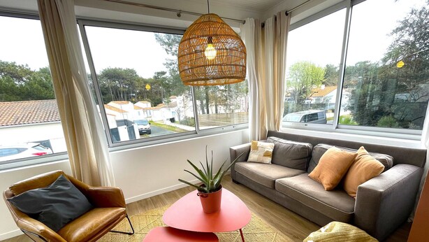 A modern living room with a leather armchair, a sofa with cushions, a small table, a potted plant, and a large window with a view of the outside. - Charming Villa Near Beach with Heated Pool, Wi-Fi, Billiards, and Garage (La Tranche-sur-Mer)