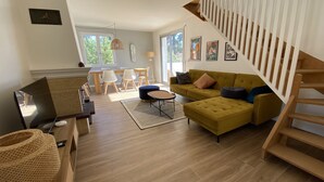A modern living room with a sofa, ottoman, and a TV. There's a staircase with a wooden railing, and a dining area with chairs and a table. - Charming Villa Near Beach with Heated Pool, Wi-Fi, Billiards, and Garage (La Tranche-sur-Mer)