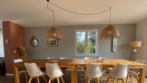 A dining area with a wooden table and white chairs, two hanging wicker lamps, a window with a view of greenery, and a floor lamp. - Charming Villa Near Beach with Heated Pool, Wi-Fi, Billiards, and Garage (La Tranche-sur-Mer)