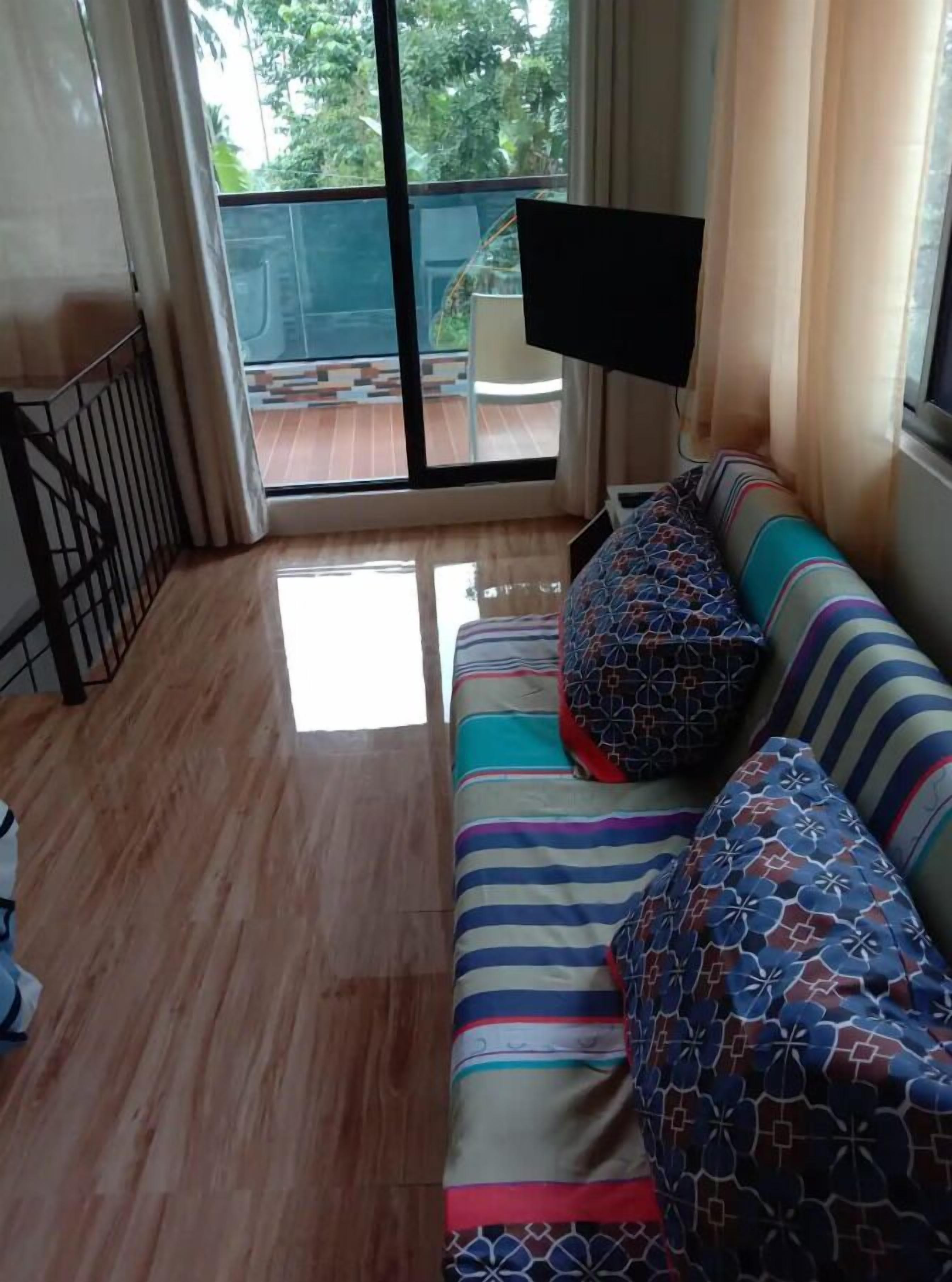 A room with a wooden floor, a striped sofa with patterned cushions, a television on a stand, and a view of a balcony with a railing.