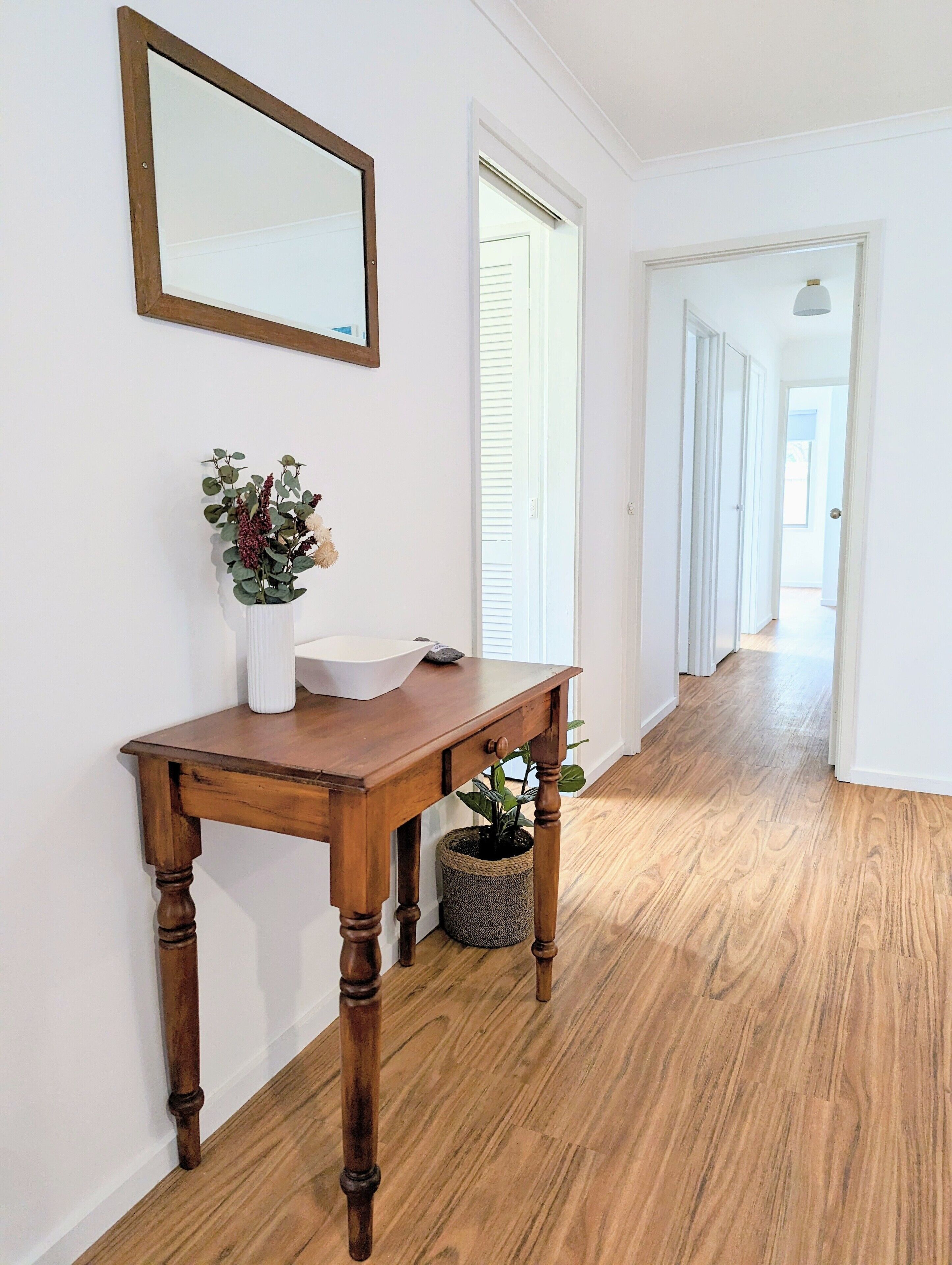 A hallway with a wooden floor, a white wall, a wooden console table with a vase of flowers, and a mirror.
