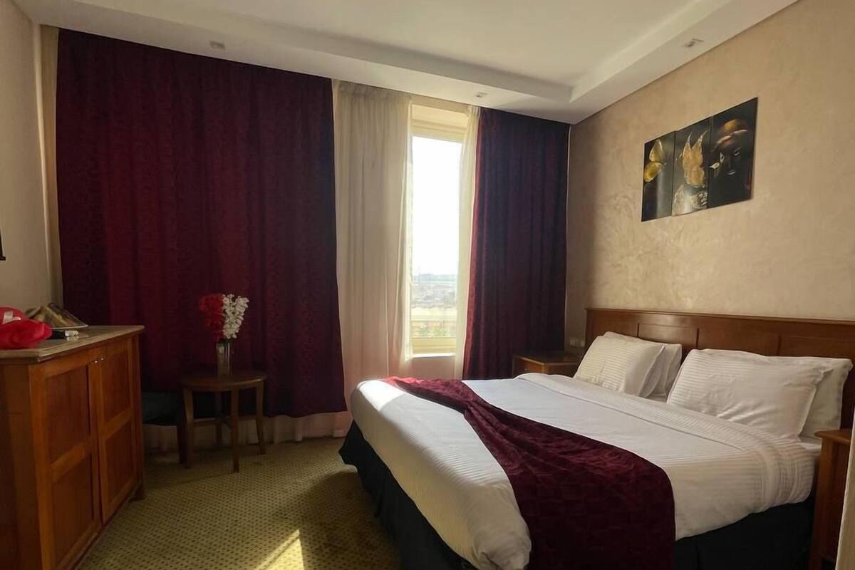 A hotel room with a bed, a desk, a chair, a window with curtains, and a painting on the wall.