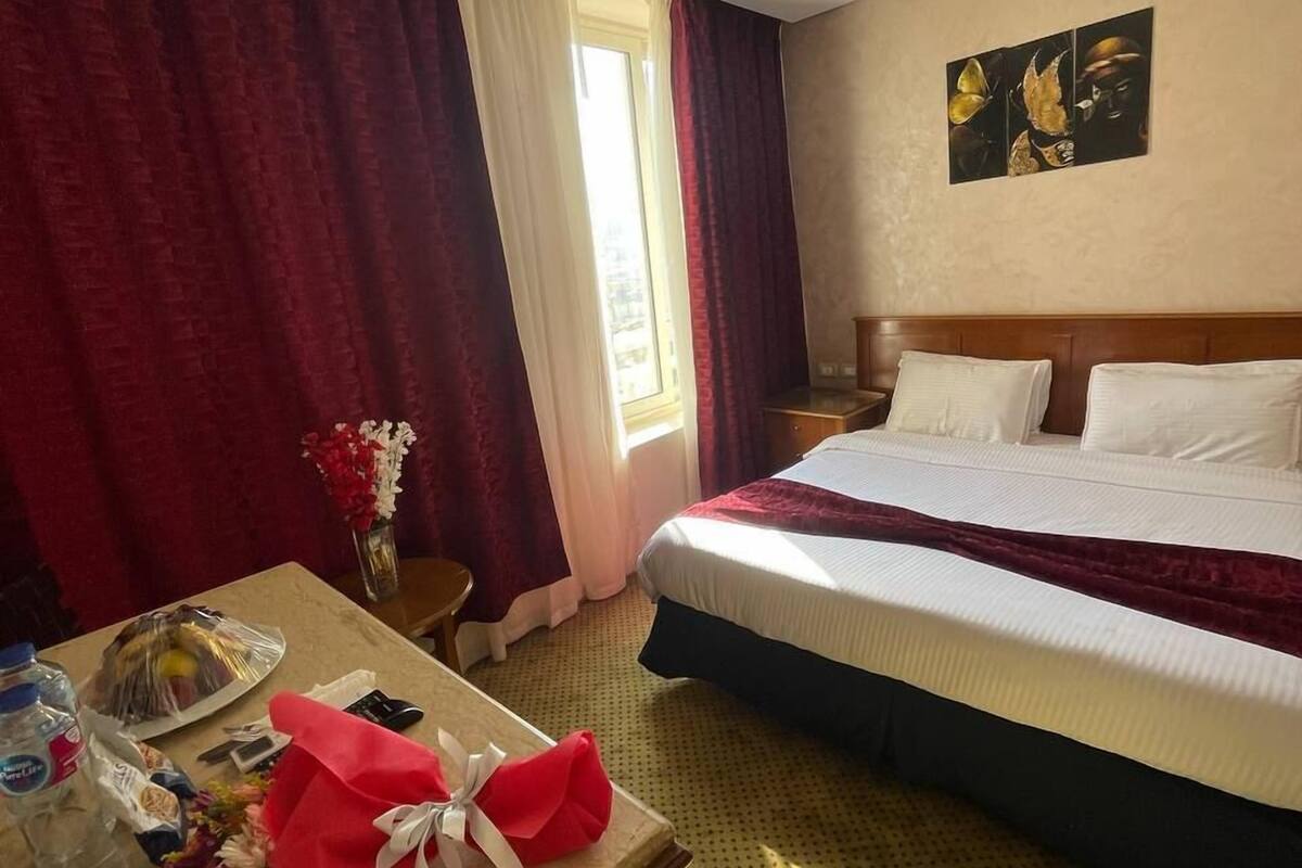 A hotel room with a bed, a desk with a vase of flowers, a table with a red gift bag, and a window with curtains.