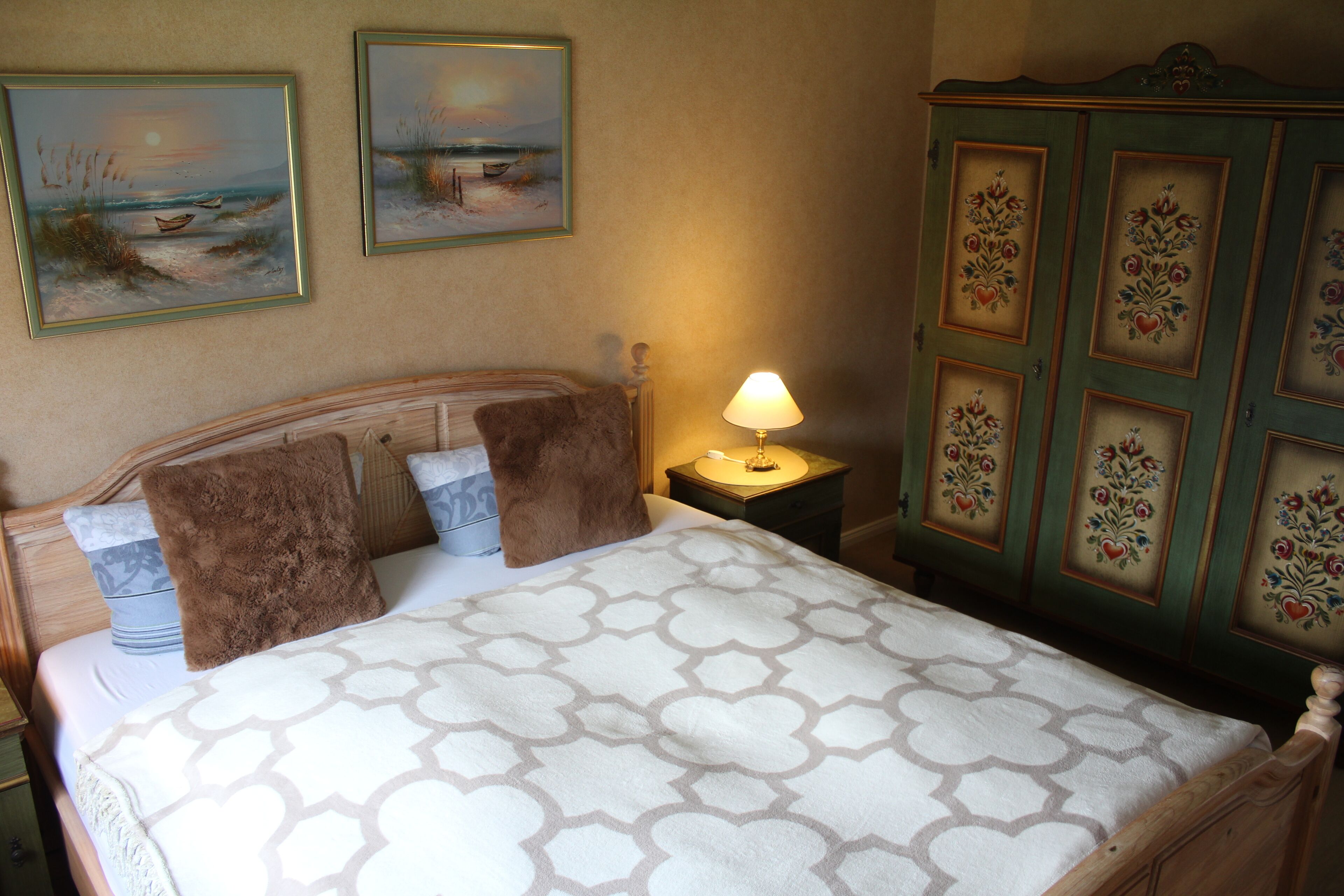 A neatly made bed with a patterned bedspread, a wooden headboard, and two pillows. There is a nightstand with a lamp and a decorative cabinet with floral designs.