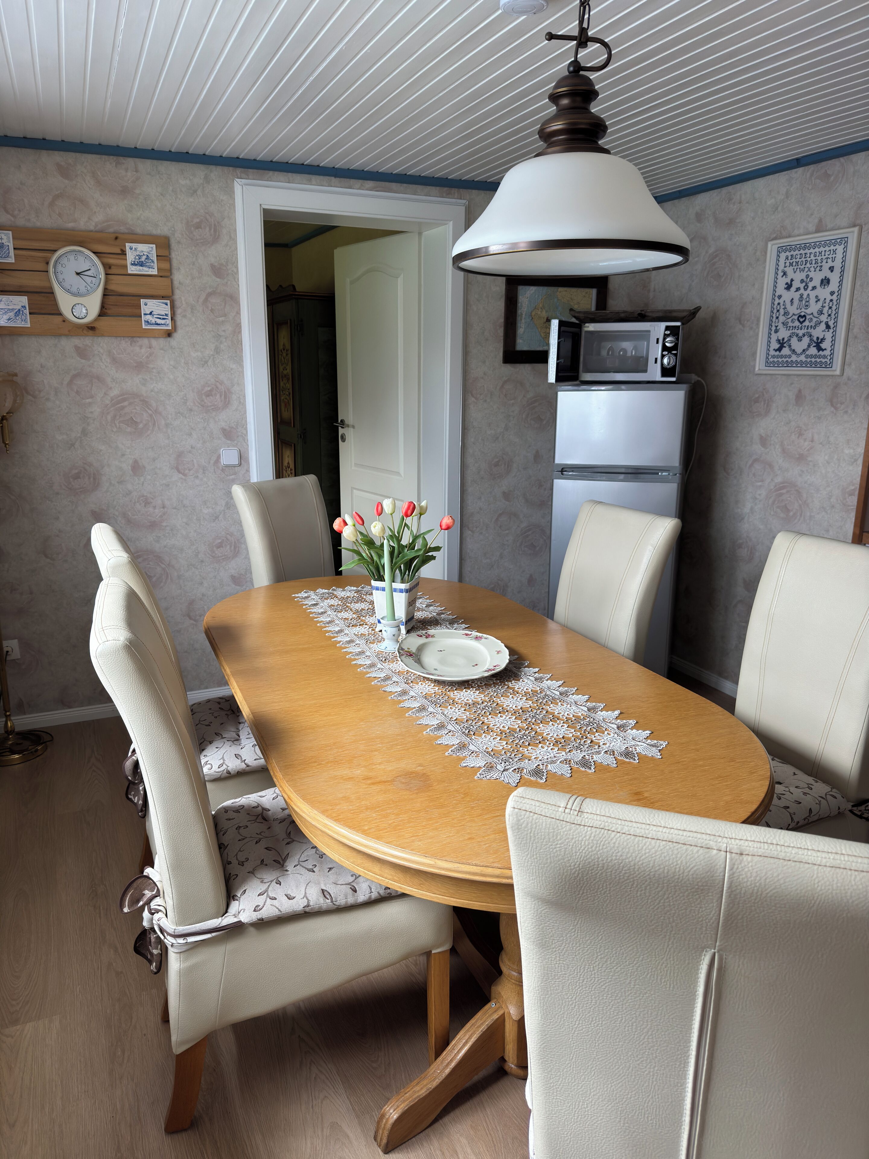 Ein Essbereich mit einem Holztisch, weißen Polsterstühlen, einer Tischläufer aus Spitze, einer Vase mit Blumen, einer Uhr, einer Mikrowelle und einem Kühlschrank.