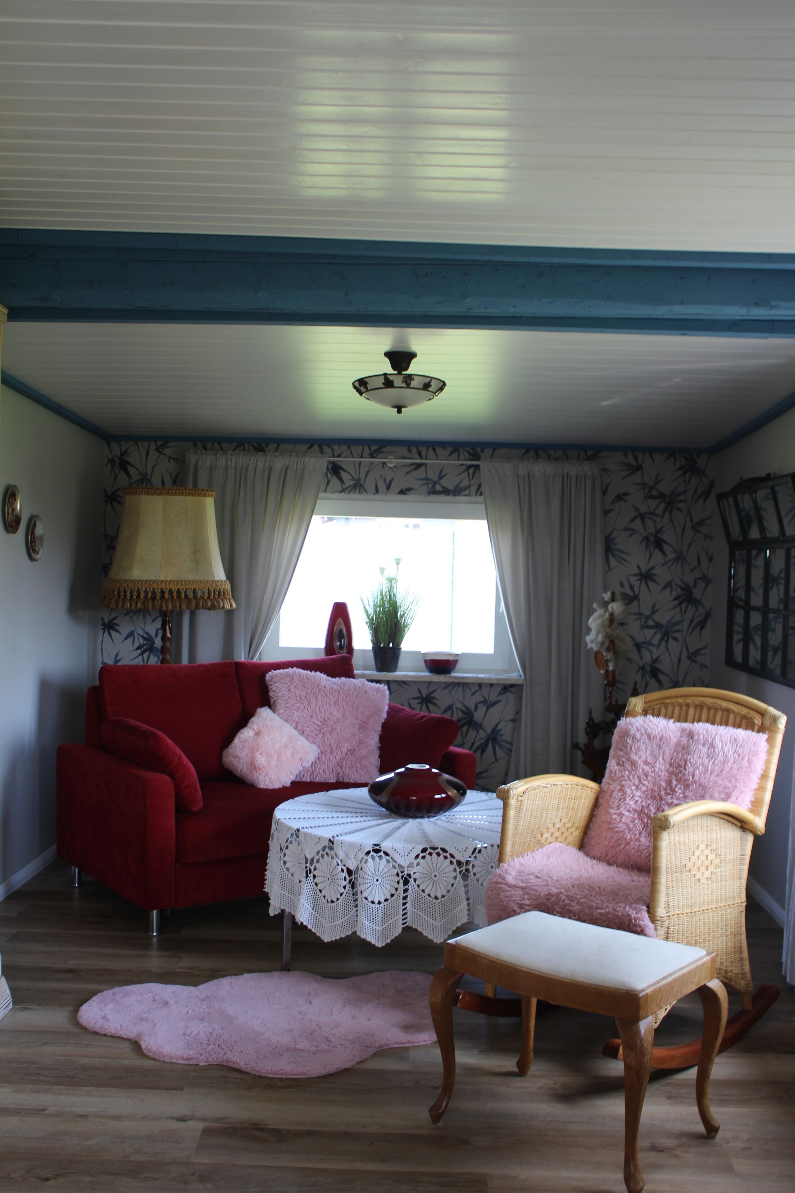 A room with a red sofa, a wicker chair, a small table with a lace tablecloth, and a pink rug.