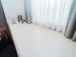 A room with a white bed, a wooden chair, a small table with a glass container, and sheer curtains.