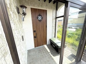 A wooden door with a decorative glass panel, a wall-mounted light fixture, and a view of greenery through a window. - Higashikawa HomeHome base for AsahikawaBiei  - higashikawa home / Kamikawagun Hokkaidō (Kamikawagun)