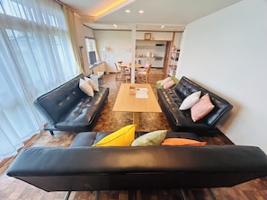 A modern living room with two black leather sofas, a wooden coffee table, and a dining area with a kitchen in the background. - Higashikawa HomeHome base for AsahikawaBiei  - higashikawa home / Kamikawagun Hokkaidō (Kamikawagun)