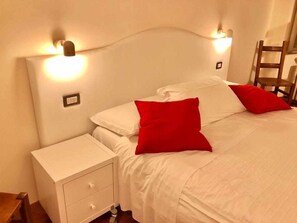 Una cama bien hecha con ropa blanca y almohadas rojas, una mesita de noche con cajones e iluminación en la pared.