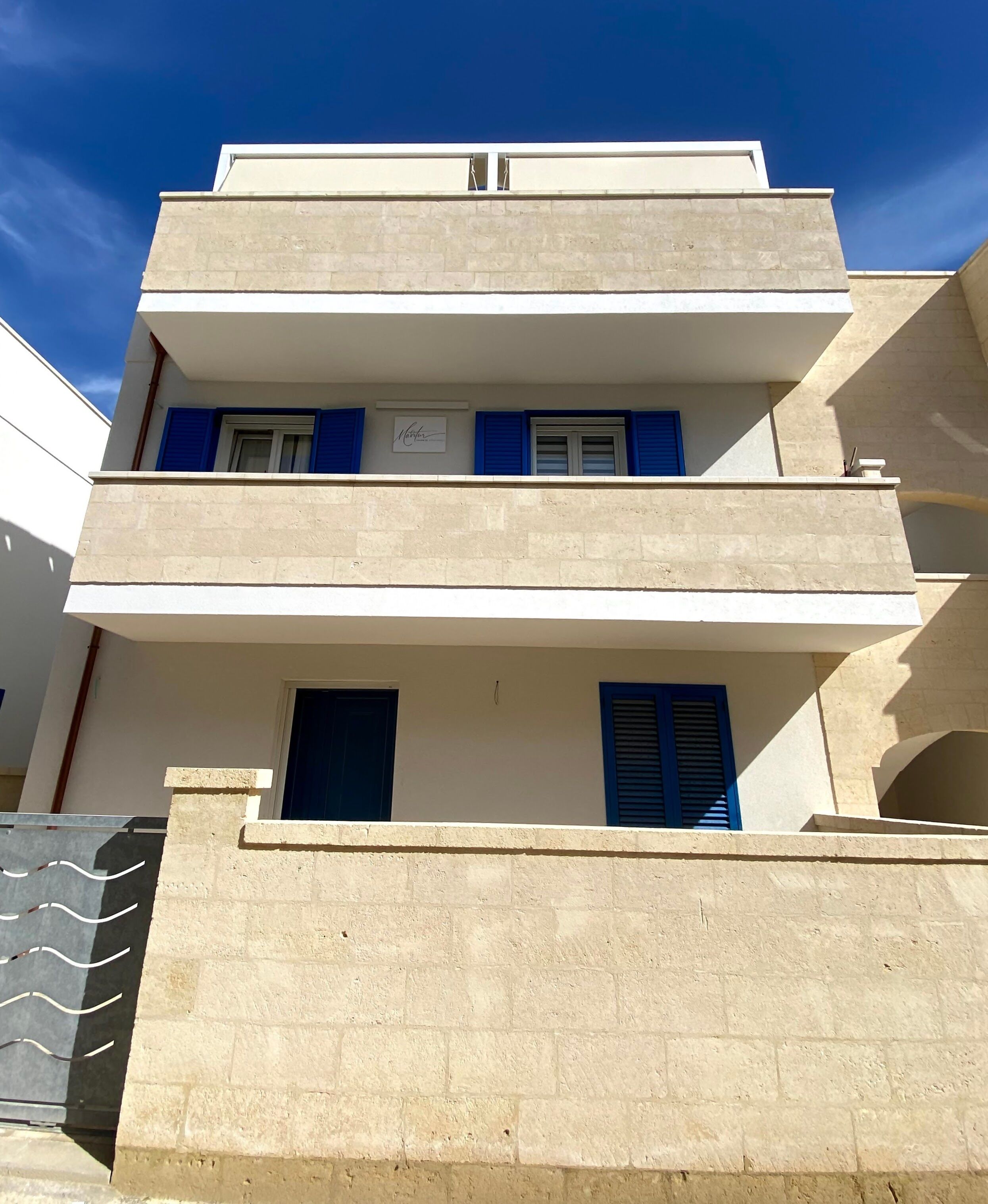 A multi-story building with a flat facade, blue shutters on windows, and a balcony on the second floor.