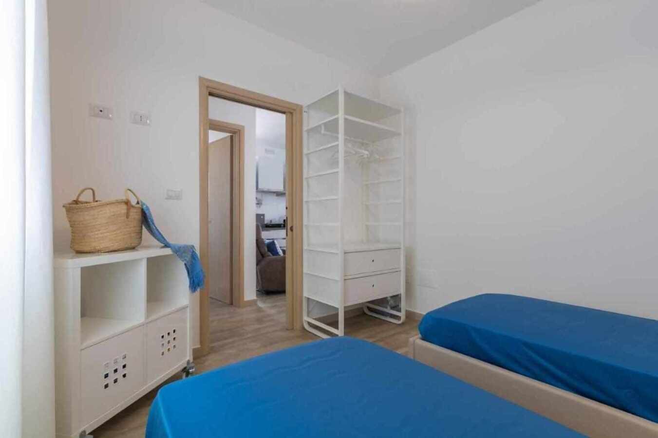 A small, clean room with two beds, a wardrobe, and a shelf with a basket.