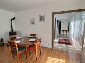 A dining area with a round wooden table, four chairs, and a black wood-burning stove. There are two framed pictures on the wall.