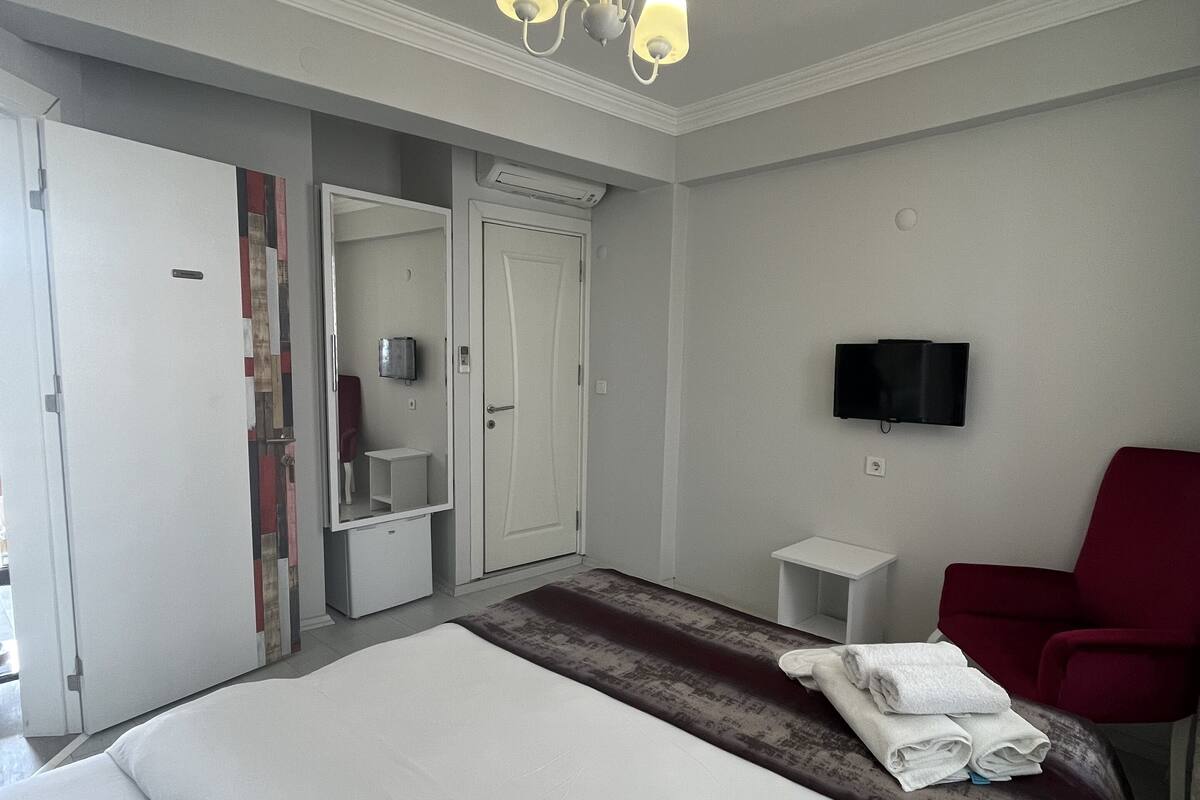 A hotel room with a large bed, a red armchair, a TV mounted on the wall, and a mirror above the door.
