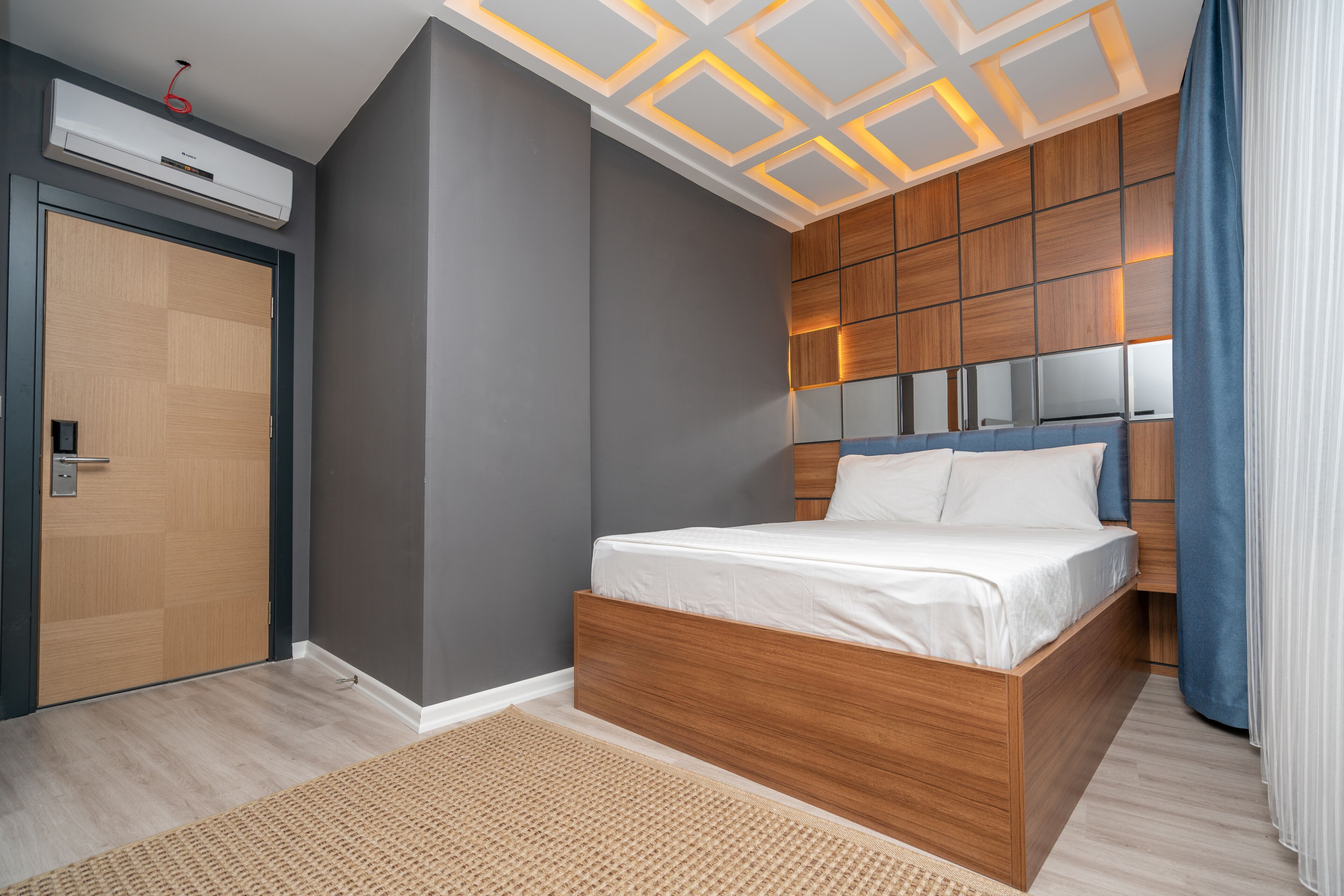 A modern bedroom with a wooden bed, grey walls, and a ceiling with geometric lighting.