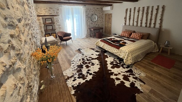 Une chambre avec un mur en pierre, des poutres apparentes, un lit recouvert d’un tapis en peau de vache, un bureau en bois et une chaise.